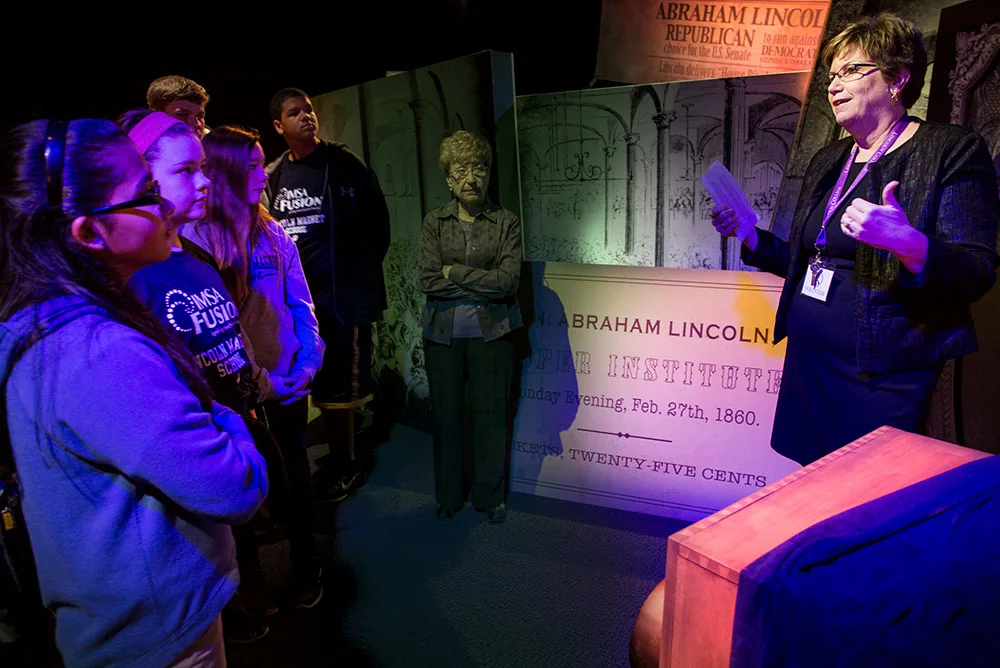 A museum docent leading a group of visitors through a lit exhibit space.
