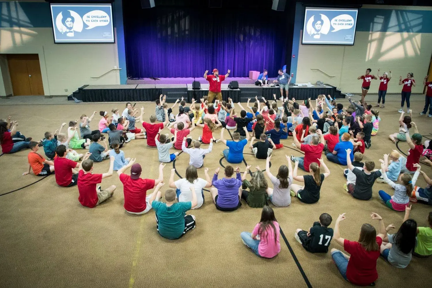 A large group of schoolchildren seated on the floor of an auditorium during a program.