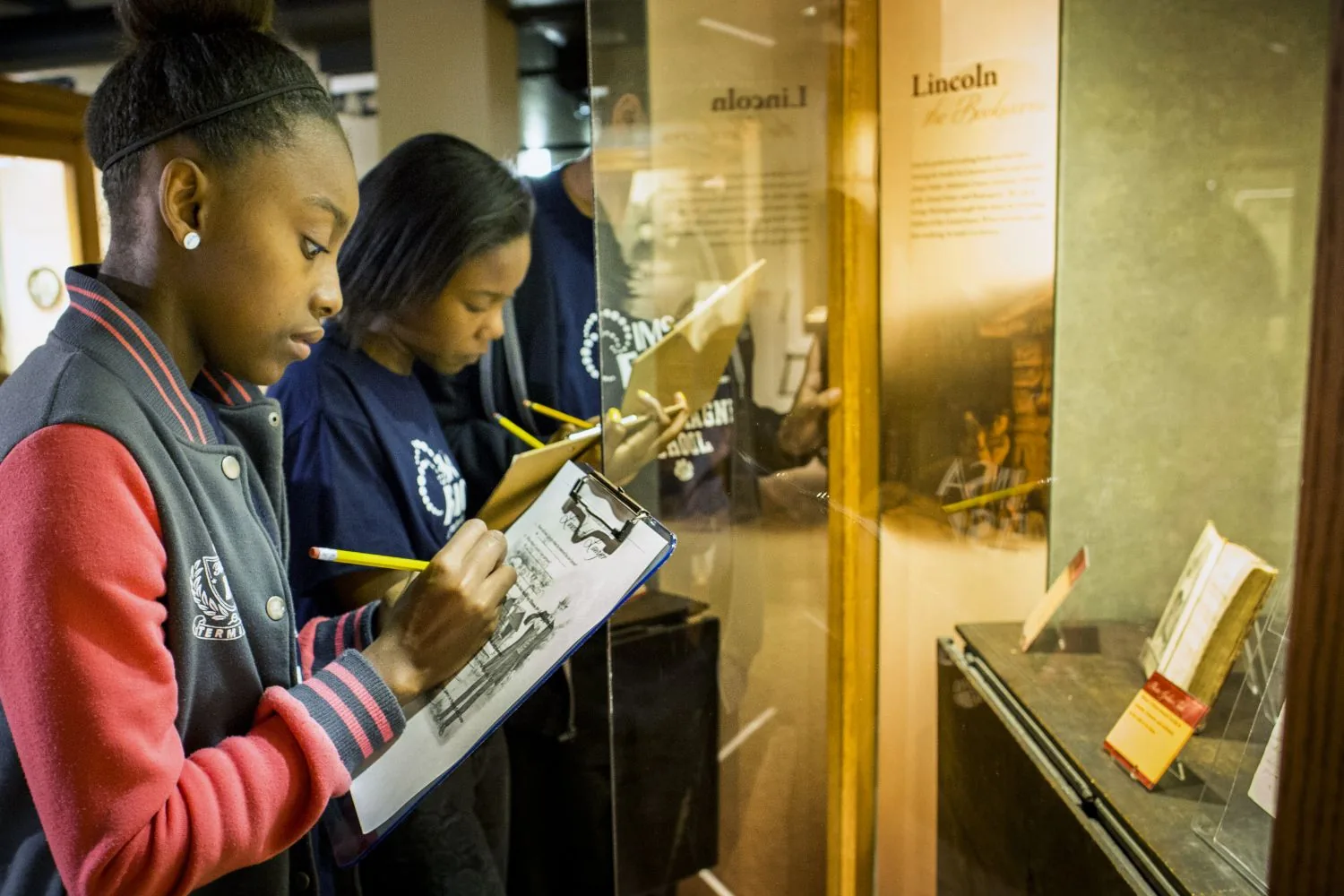 Two students with clipboards examining a gallery case during a school field trip.
